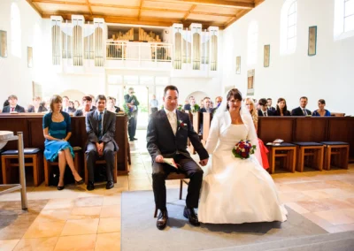 Brautpaar sitzt bei der Trauung in der Christuskirche Gauting, umgeben von Gaesten, Hochzeitsfotografie nahe Muenchen.