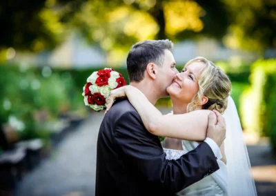 Brautpaar umarmt sich im Hofgarten Muenchen, rote Rosen im Brautstrauss, Hochzeitsfoto bei sonnigem Wetter.