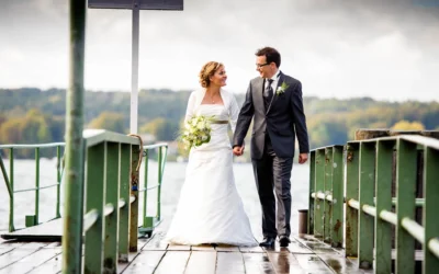 Ein Brautpaar geht lächelnd Hand in Hand auf einem Bootssteg. Die Braut trägt ein weißes Kleid mit Blumenstrauß.