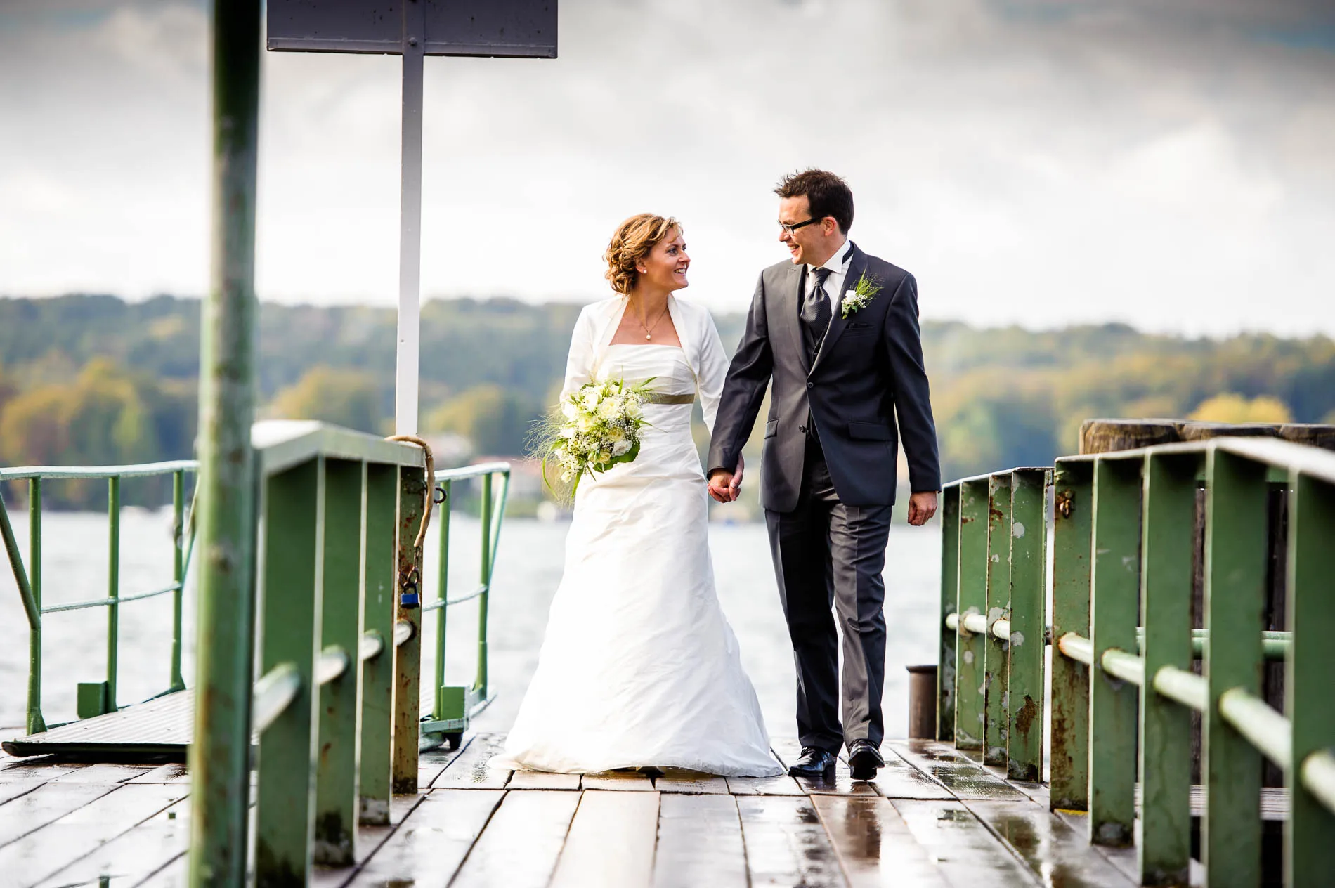 Brautpaar geht Hand in Hand auf einem Steg am Starnberger See, Hochzeitsportrait mit See im Hintergrund.