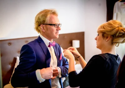 Bräutigam in blauem Anzug wird beim Getting Ready in Landshut von einer Frau angepasst. Hochzeitsfoto.