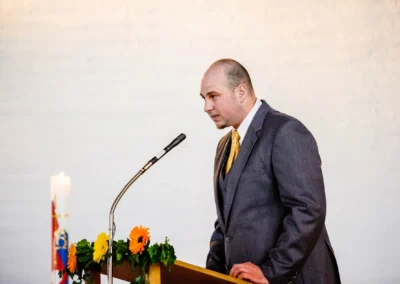 Mann im Anzug spricht am Mikrofon, Blumenschmuck am Pult. Kirchliche Trauung nordlich von Muenchen.