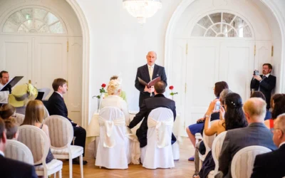 Paar sitzt bei Trauung, dekoriert mit Rosen. Redner mit Buch, Gäste fotografieren. Elegantes Ambiente, mit großen weißen Türen.