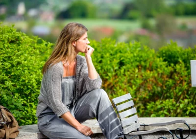 Frau im grauen Kleid sitzt nachdenklich auf einer Bank im Aschbacher Hof, umgeben von gruenen Bueschen.