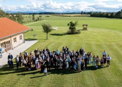 Hochzeitsfoto einer großen Gruppe auf einer Wiese bei Archehof Schlickenrieder in Otterfing, aufgenommen aus der Vogelperspektive.