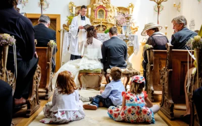 Brautpaar kniet in einer Kirche vor dem Altar, Priester liest. Kinder sitzen am Boden, Gäste auf Holzbänken, festlich gekleidet.