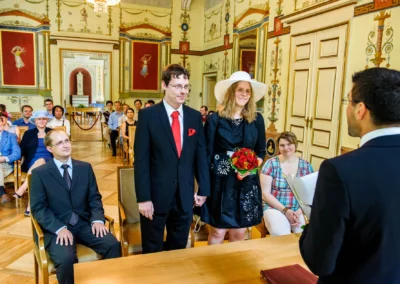 Brautpaar bei standesamtlicher Trauung im Schloss Ismaning, umgeben von Gästen und Verzierungen im Raum.