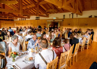 Gäste sitzen in Holzsaal bei einer Hochzeit nahe Otterfing, festlich gedeckte Tische, entspannte Atmosphäre.