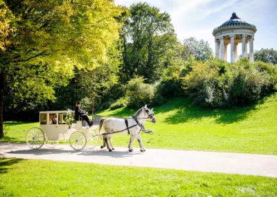 Weiße Kutsche mit Brautpaar im englischen Garten Schwabing, Hochzeitsshooting unter sonnigem Himmel, grüne Landschaft.