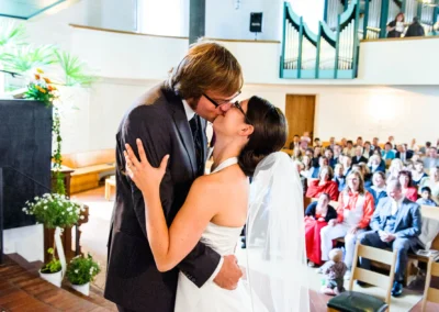 Brautpaar beim Kuss in der Kirche, Hochzeitsfotografie bei der Trauung in der Friedenskirche Dachau, Gaeste im Hintergrund.
