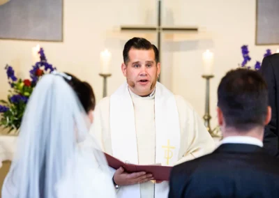 Priester spricht zu Brautpaar in Kirche bei Hochzeit in Gauting nahe Muenchen. Hochzeitsfotografie mit bunten Blumenarrangements.