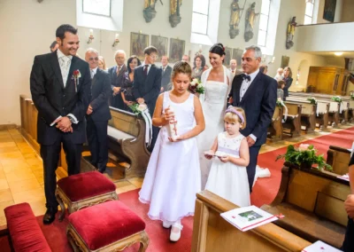 Brautpaar betritt Kirche bei kirchlicher Trauung in München. Gäste stehen auf, Mädchen tragen Kerze und Buch. Hochzeitsfoto.