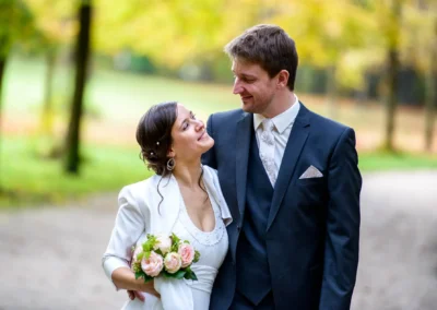 Brautpaar beim Spaziergang im englischen Garten Muenchen, sie laechelnd mit Blumenstrauss, Hochzeitsfotografie.