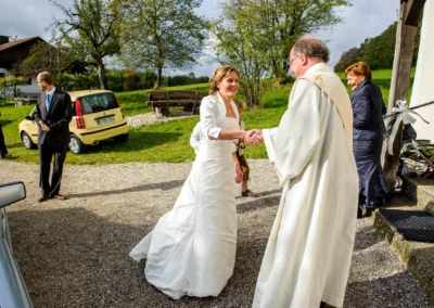 Braut begruesst Priester bei kirchlicher Trauung am Starnberger See, gruenes Auto im Hintergrund, Hochzeitsfotoreportage.