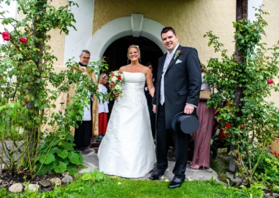 Brautpaar nach kirchlicher Trauung in Irschenberg, posiert vor der Kirche, begleitet von einem Priester.