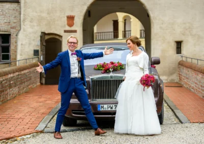 Brautpaar vor einem Auto bei Hochzeitsfotos in Landshut, Braut mit Blumenstrauss, Bräutigam in blauem Anzug.