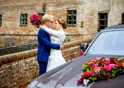 Brautpaar-Portrait bei Hochzeit auf Burg Trausnitz, Paar in Umarmung mit Brautstrauss, Auto mit Blumen im Vordergrund.