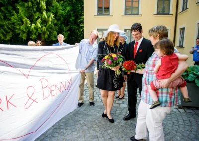 Hochzeitsfoto im Schloss Ismaning mit Brautpaar und Hochzeitsgästen, die ein großes Herzbanner halten.