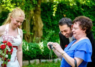Braut bei Hochzeit in Röhrmoos betrachtet mit Gästen Fotos auf Kamera, umgeben von grünem Hintergrund. Hochzeitsfoto.