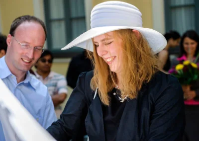 Frau mit weissem Hut lacht bei Hochzeitsfeier in Ismaning, Mann im Hintergrund lächelt. Hochzeitsfotografie nördlich von München.