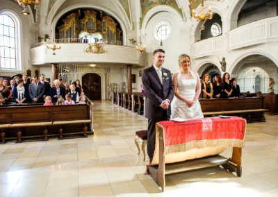Brautpaar steht bei der kirchlichen Trauung in Sankt Sylvester Muenchen, Gaeste sitzen in Kirchenbaenken.