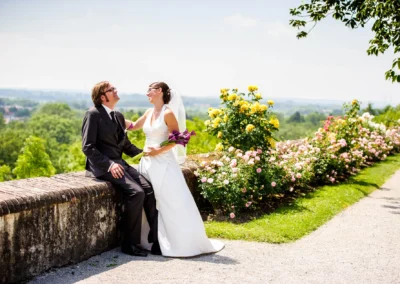 Brautpaar lacht auf einer Mauer im Hofgarten Schloss Dachau, umgeben von blühenden Rosen. Hochzeitsfotografie.