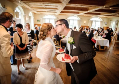 Hochzeitspaar beim Kuchenessen, umgeben von Gästen im Aschbacher Hof, festgehalten von Hochzeitsfotografen.