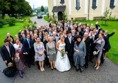 Hochzeitsgesellschaft vor Wallfahrtskirche Wilparting, Brautpaar lacht umgeben von Gaesten im Freien.