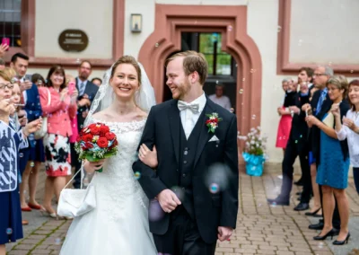 Brautpaar laechelnd vor Kirche mit Gaesten bei Hochzeitsreportage in Sankt Nikolaus Schalkhausen-Ansbach.