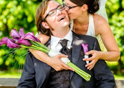 Brautpaar auf Bank im Hofgarten Schloss Dachau, lachend und vertraut, mit lila Blumenstrauss. Hochzeitsfotografie.