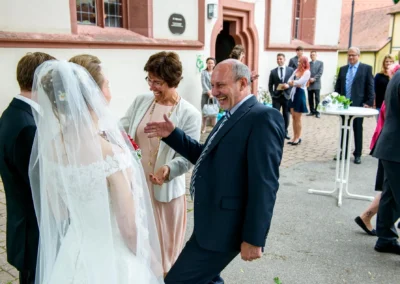 Braut in Spitzenkleid mit Schleier lacht mit Gaesten bei einer Hochzeit in Ansbach vor einer Kirche draußen.