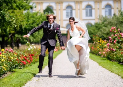 Bräutigam und Braut laufen Hand in Hand im Hofgarten Schloss Dachau, umgeben von bunten Blumen. Hochzeitsfoto.