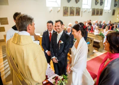 Brautpaar bei kirchlicher Trauung in München, lächelnd vor dem Priester, Gäste im Hintergrund. Hochzeitsfoto.