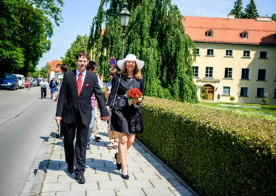 Paar spaziert Hand in Hand bei Hochzeit in Ismaning, Frau hält Blumenstrauss, Schloss im Hintergrund.