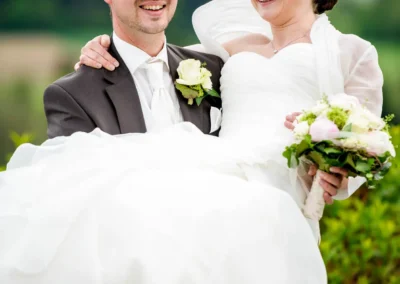 Brautpaar lacht bei Hochzeitsfoto im Aschbacher Hof, sie in weissem Kleid mit Blumen, er in Anzug mit Rose.