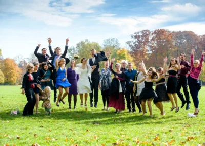 Hochzeitsgesellschaft im englischen Garten in München, springende Menschen, festliche Kleidung, sonniger Tag.