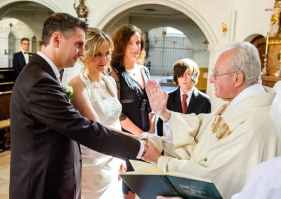 Priester leitet kirchliche Trauung in Sankt Sylvester Muenchen, Brautpaar und Gaeste im Hintergrund, Hochzeitsfotografie.