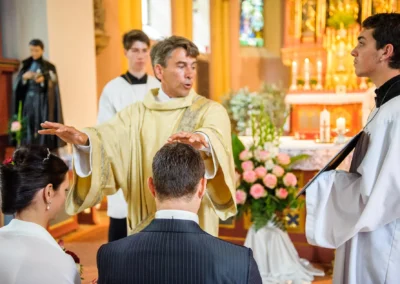 Priester segnet Brautpaar bei kirchlicher Trauung in München. Kerzen und Blumen im Hintergrund betonen das Ambiente.