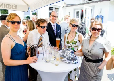Gäste mit Sonnenbrillen bei Hochzeitsfeier in Markt Indersdorf, lachend am Tisch unter Sonnenschirm.