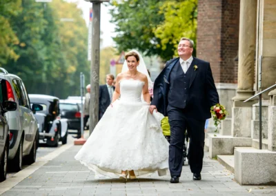 Brautpaar bei Hochzeitsreportage auf Gehweg in Muenchen-Lehel, Braut im weissen Kleid, Bräutigam mit Blumenstrauss.