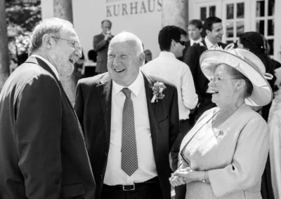 Hochzeit in Bad Toelz: Drei Gaeste im Gespraech vor dem Kurhaus, festlich gekleidet und froehlich im Freien.