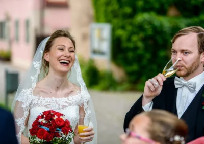Braut lacht mit Blumenstrauss in Ansbach, Hochzeitsfotografie; daneben Bräutigam mit Sektglas.