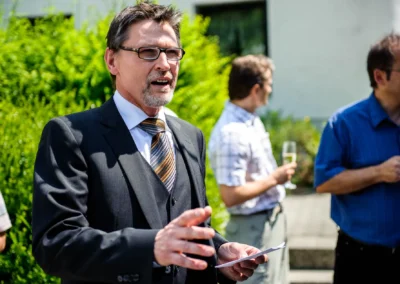 Mann in Anzug bei Hochzeitsfeier in Ismaning, spricht vor Gästen im Freien. Teil einer Hochzeitsreportage.