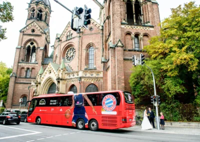 Brautpaar in Lehel vor rotem Bus und Kirche, festgehalten auf einem Hochzeitsfoto in Muenchen naehe Isar.