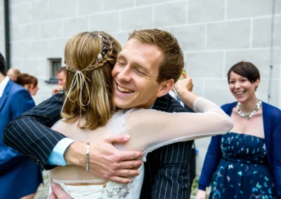 Umarmung eines Paars bei Hochzeit im Schloss Gruenau, Hochzeitsreportage zeigt froehliche Gaeste im Hintergrund.