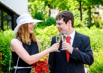 Paar stößt mit Sektgläsern an, Frau mit Hut und Blumenstrauß, Hochzeit nördlich von München im Grünen.