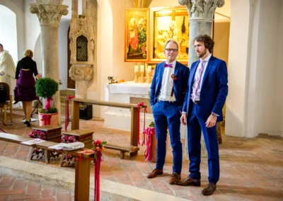Zwei Maenner in blauen Anzuegen stehen in einer Kirche bei der Hochzeit in Burg Trausnitz vor einem Altar.