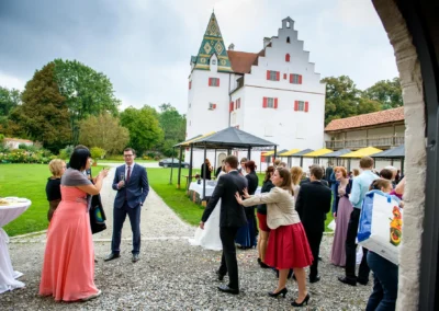 Gäste bei einer Hochzeitsfeier im Schloss Grünau, elegant gekleidet, sonniger Tag, grüne Wiese im Hintergrund.