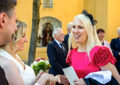 Braut mit Blumenstrauss bei der Hochzeit in Schwabing, Muenchen, laechelt mit Gaesten in bunten Kleidern im Hintergrund.