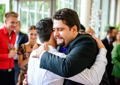 Umarmung zweier Maenner bei einer Hochzeit im Palmenhaus Schloss Nymphenburg, Gaeste im Hintergrund, Hochzeitsreportage.